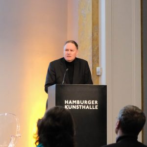Hamburgs Senator für Kultur und Medien eröffnet mit einer Rede die Veranstaltung „60 Jahre Besuchsprogramm für jüdische ehemalige Hamburger Bürger und Bürgerinnen“. Er steht hinter einem Rednerpult mit der Aufschrift „Hamburger Kunsthalle“. Im Vordergrund sind man die Köpfe zweier Personen aus dem Publikum.