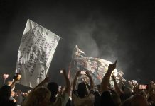 Deichkind-Mitgründer Malte Pittner verstorben Ein Konzertbild, auf dem eine Menschenmenge vorne steht. Im Hintergrund weht eine riesige weiße Fahne mit Text. Auf der Bühne steht eine große Röhre, auf der jemand kniet.