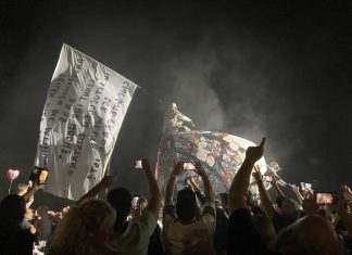 Deichkind-Mitgründer Malte Pittner verstorben Ein Konzertbild, auf dem eine Menschenmenge vorne steht. Im Hintergrund weht eine riesige weiße Fahne mit Text. Auf der Bühne steht eine große Röhre, auf der jemand kniet.