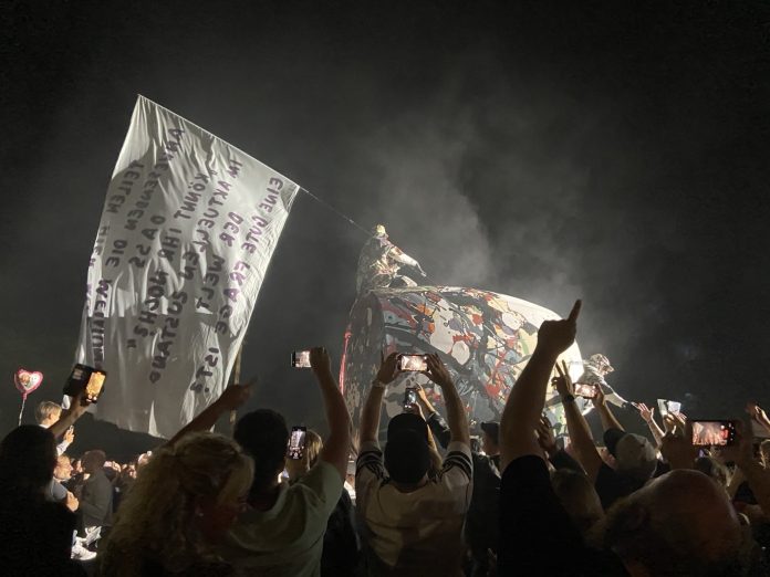 Deichkind 2024 Ein Konzertbild, auf dem eine Menschenmenge vorne steht. Im Hintergrund weht eine riesige weiße Fahne mit Text. Auf der Bühne steht eine große Röhre, auf der jemand kniet.