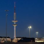 Hamburger Fernsehturm soll 2031 öffnen Der Hamburger Fernsehturm über den Dächern der Stadt vor dunklem Himmel. Neben dem Fernsehturm sieht man große Scheinwerfer.