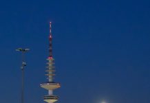 Hamburger Fernsehturm soll 2031 öffnen Der Hamburger Fernsehturm über den Dächern der Stadt vor dunklem Himmel. Neben dem Fernsehturm sieht man große Scheinwerfer.