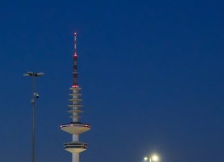 Hamburger Fernsehturm soll 2031 öffnen Der Hamburger Fernsehturm über den Dächern der Stadt vor dunklem Himmel. Neben dem Fernsehturm sieht man große Scheinwerfer.