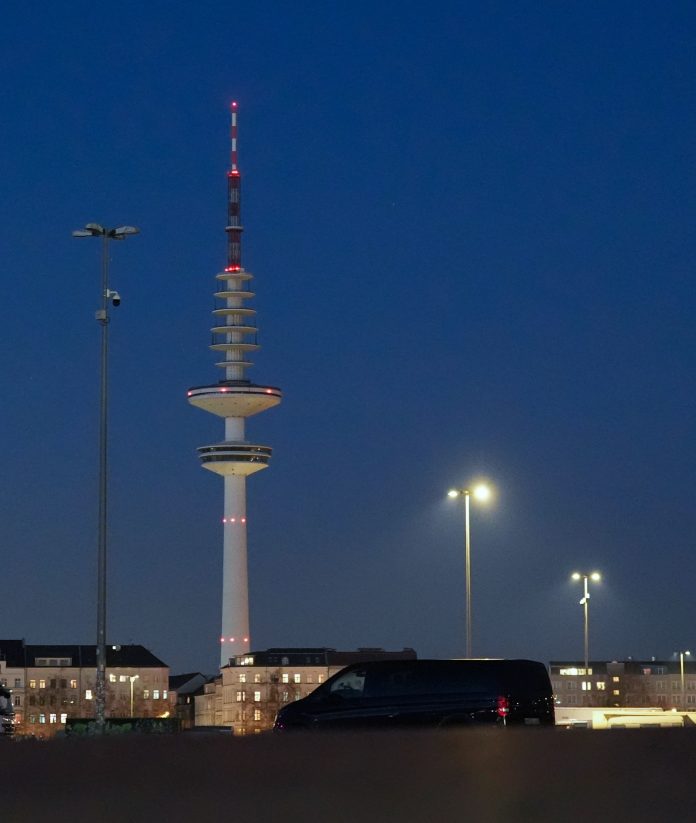 Fernsehturm Heinrich-Hertz-Turm Der Hamburger Fernsehturm über den Dächern der Stadt vor dunklem Himmel. Neben dem Fernsehturm sieht man große Scheinwerfer.