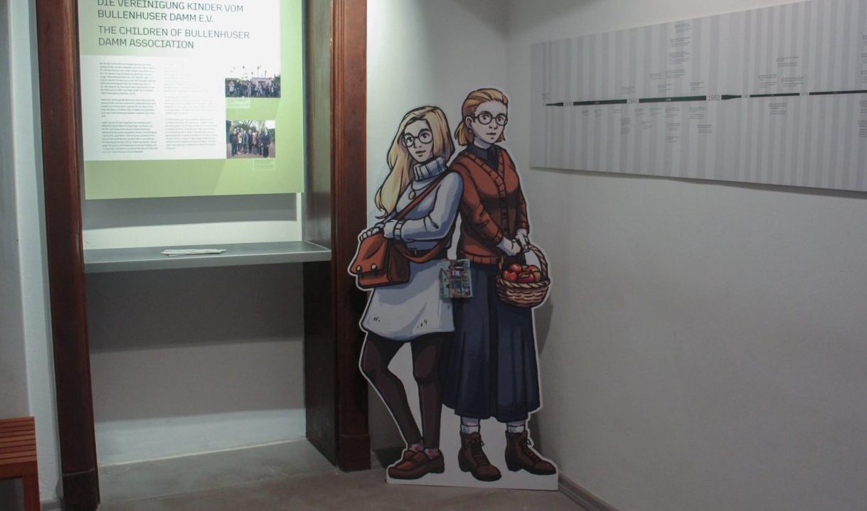 Ein Museumsraum mit einer Zeitleiste an der Wand. In der Ecke steht ein Aufsteller von zwei lebensgroßen gezeichneten Frauen, die Rücken an Rücken beieinander stehen.