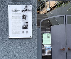 Es ist eine Gedenktafel an einem Grundstückseingang mit der Überschrift „Ein Haus mit bewegter Geschichte“ zu sehen. Daneben hängt ein laminiertes Plakat mit einem alten Foto und unleserlichen Text und ein transparanter Kasten mit Infomaterial zum Mitnehmen. Das Haus befindet sich am Kahlkamp 1A in Hamburg Blankenese.