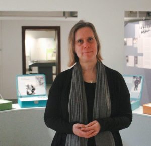 Eine Frau mit blonden Haaren steht mit gefalteten Händen in einem Raum. Im Hintergrund sind bunte Metallkoffer zu sehen, in denen die Informationen zu den Verstorbenen ausgestellt sind.