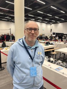 Ein Mann Mitte 50, der im Portrait fotografiert ist und vor seiner Kugelbahn steht.