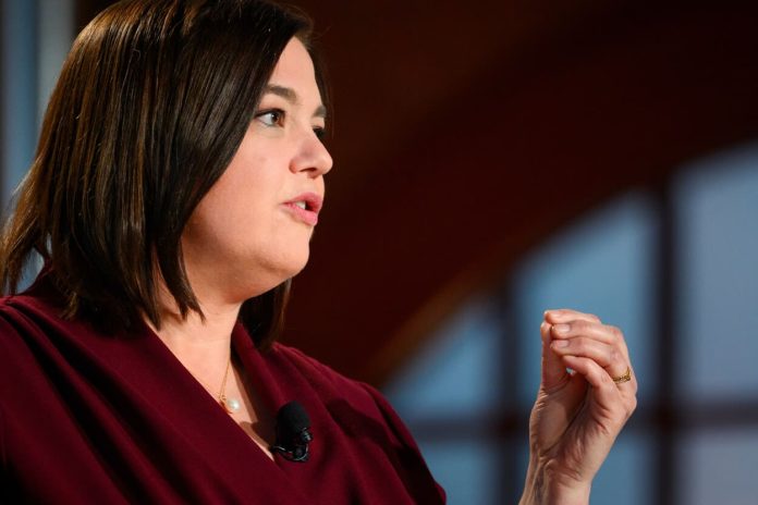 Das Bild zeigt eine Frau mit dunklen Haaren und einer roten Bluse. Sie befindet sich auf der linken Seite des Bildes, schaut nach rechts und ist von der Seite fotografiert.