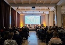 Auf der Suche nach jüdischen Familiengeschichten Blick von hinten aus dem Publikum auf ein Podium mit sechs sitzenden Personen vor dem Schriftzug „60 Jahre Besuchsprogramm für jüdische ehemalige Hamburger Bürger und Bürgerinnen“