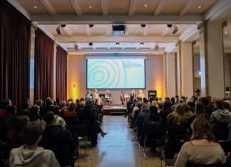 Auf der Suche nach jüdischen Familiengeschichten Blick von hinten aus dem Publikum auf ein Podium mit sechs sitzenden Personen vor dem Schriftzug „60 Jahre Besuchsprogramm für jüdische ehemalige Hamburger Bürger und Bürgerinnen“