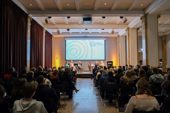Podiumsdiskussion 60 Jahre Besuchsprogramm Blick von hinten aus dem Publikum auf ein Podium mit sechs sitzenden Personen vor dem Schriftzug „60 Jahre Besuchsprogramm für jüdische ehemalige Hamburger Bürger und Bürgerinnen“