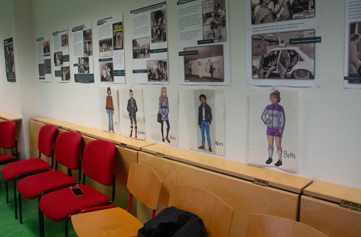 Ein Raum mit roten Stühlen und heller Holzverkleidung. An den Wänden hängen Poster zu rechter Gewalt in Hamburg. Vier gezeichnete Charaktere des Videospiels "Erinnern. Die Kinder vom Bullenhuser Damm” hängen ausgedruckt auf DIN A4 Blättern an der Wand.