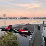 Zwei Verletzte durch Ruderunfall Das Bild steigt einen Steg auf der Außenalster, an dem zwei Motorboote festgemacht sind. Hinter dem Zaun, der den Steg absperrt sind zwei Polizisten zu sehen. Im Hintergrund sieht man den Fernsehturm und am Himmel Farben vom Sonnenaufgang.
