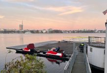 Zwei Verletzte durch Ruderunfall Das Bild steigt einen Steg auf der Außenalster, an dem zwei Motorboote festgemacht sind. Hinter dem Zaun, der den Steg absperrt sind zwei Polizisten zu sehen. Im Hintergrund sieht man den Fernsehturm und am Himmel Farben vom Sonnenaufgang.