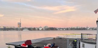 Zwei Verletzte durch Ruderunfall Das Bild steigt einen Steg auf der Außenalster, an dem zwei Motorboote festgemacht sind. Hinter dem Zaun, der den Steg absperrt sind zwei Polizisten zu sehen. Im Hintergrund sieht man den Fernsehturm und am Himmel Farben vom Sonnenaufgang.