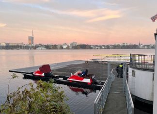 Zwei Verletzte durch Ruderunfall Das Bild steigt einen Steg auf der Außenalster, an dem zwei Motorboote festgemacht sind. Hinter dem Zaun, der den Steg absperrt sind zwei Polizisten zu sehen. Im Hintergrund sieht man den Fernsehturm und am Himmel Farben vom Sonnenaufgang.