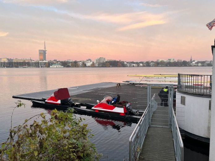 Ruderboot Außenalster Das Bild steigt einen Steg auf der Außenalster, an dem zwei Motorboote festgemacht sind. Hinter dem Zaun, der den Steg absperrt sind zwei Polizisten zu sehen. Im Hintergrund sieht man den Fernsehturm und am Himmel Farben vom Sonnenaufgang.
