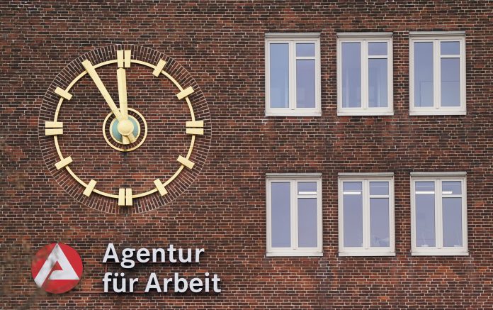 Außenansicht auf ein Backsteingebäude mit einer goldenen Uhr und dem roten Logo der Agentur für Arbeit.
