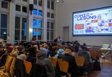 Comic-Lesung: Mehr als bunte Kästchen Das Bild zeigt einen Saal in dem rund 70 Personen in mehreren Stuhlreihen auf Holzstühlen vor einer Leinwand sitzen. Auf der Leinwand wird das Veranstaltungsposter der Comic-Lesung angezeigt.