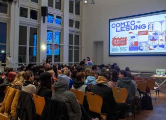 Comic-Lesung: Mehr als bunte Kästchen Das Bild zeigt einen Saal in dem rund 70 Personen in mehreren Stuhlreihen auf Holzstühlen vor einer Leinwand sitzen. Auf der Leinwand wird das Veranstaltungsposter der Comic-Lesung angezeigt.