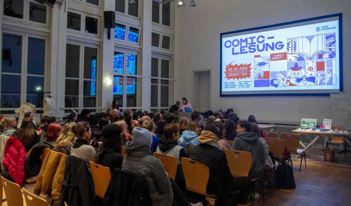 Das Bild zeigt einen Saal in dem rund 70 Personen in mehreren Stuhlreihen auf Holzstühlen vor einer Leinwand sitzen. Auf der Leinwand wird das Veranstaltungsposter der Comic-Lesung angezeigt.