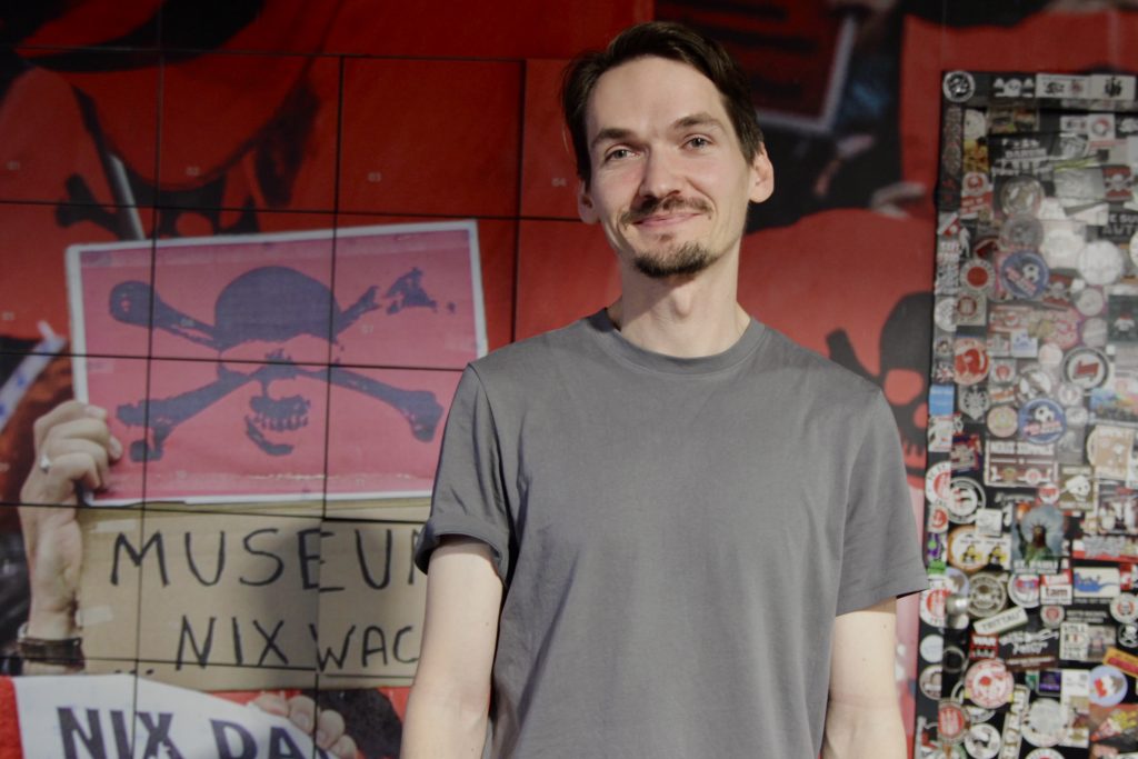 Ein Mann lächelt in die Kamera. Im Hintergrund eine rote Wand mit dem FC St. Pauli Totenkopf. Rechts davon eine Tür, die mit vielen Stickern beklebt ist.