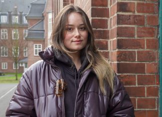 „Ich möchte einfach keine Mutter sein“ Eine junge Frau schaut in die Kamera. Sie hat lange braune Haare und steht vor einer Mauer. Links von ihr ist ein Innenhof im Hintergrund zu erkennen.