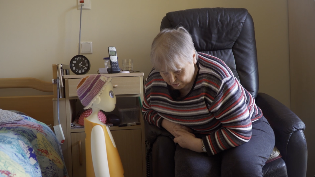 Ein KI-Roboter unterhält sich mit einer Bewohnerin eines Alten-und Pflegeheims. Die Bewohnerin sitzt auf einem schwarzen Ledersessel, der Roboter steht neben ihrem Bett.