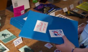 Auf dem Bild sieht man die Hände einer Person, die ein blaues Comic Heft hält. Darauf steht in roter Schreibschrift My Preschool Romance. Im Hintergrund sieht man einen Tisch auf dem viele andere Comic Hefte liegen.