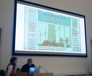 Auf dem Bild ist eine Leinwand zu sehen, auf der das Titelblatt einer illustrierten Tageszeitung zu sehen ist. Die Illustration ist in Grün-Tönen gehalten. Vor der Leinwand steht ein Mann in schwarzer Jacke und grüner Schildkappe. Er steht an einem Pult und schaut auf einen Laptop. Links neben ihm steht eine Frau mit langen, braunen Haaren. Die trägt ein hellblaues Hemd und einen rot-blau-gelb gestreiften Strickpullunder.