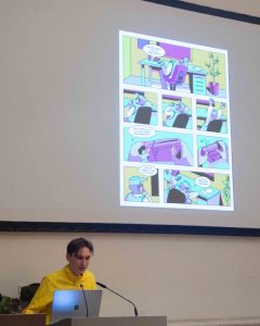 Ein Mann mit braunen, kurzen Haaren und gelber Jacke steht an einem Pult und spricht in ein Mikrofon. Hinter ihm kann man auf einer Leinwand einen bunten Comic sehen.