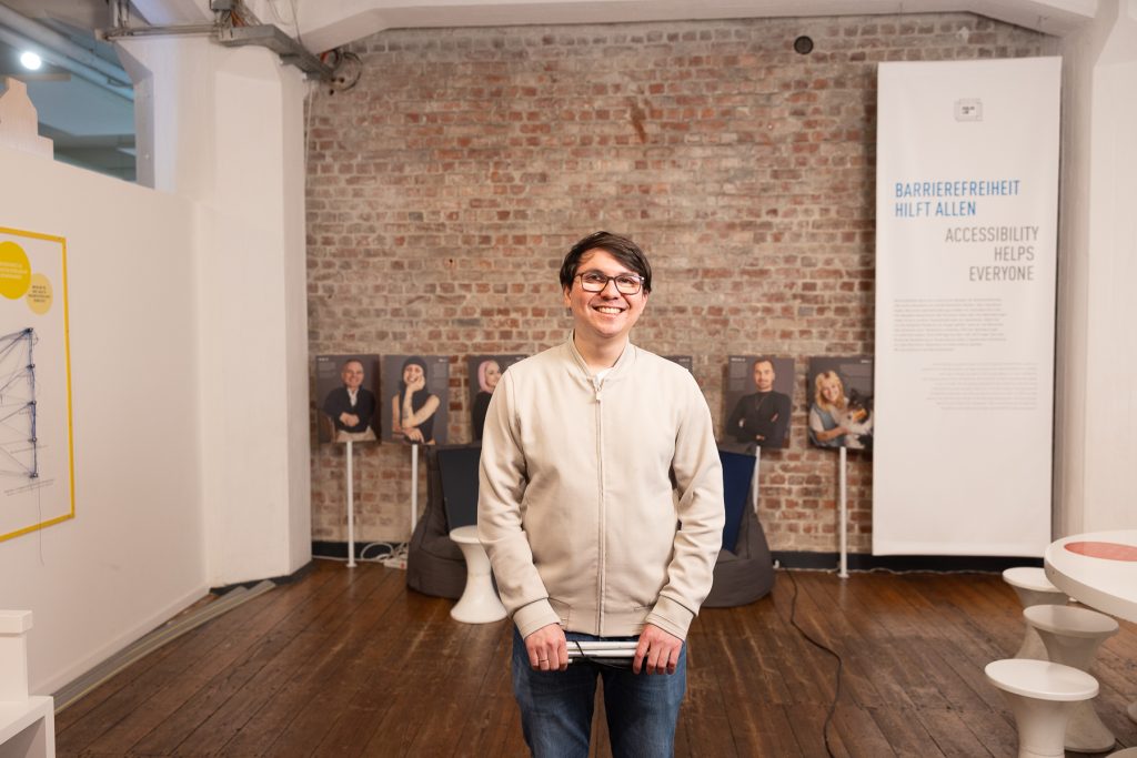 Ein junger Mann mit einem Lagstock in den Händen lächelt in die Kamera. Er trägt eine Brille.
