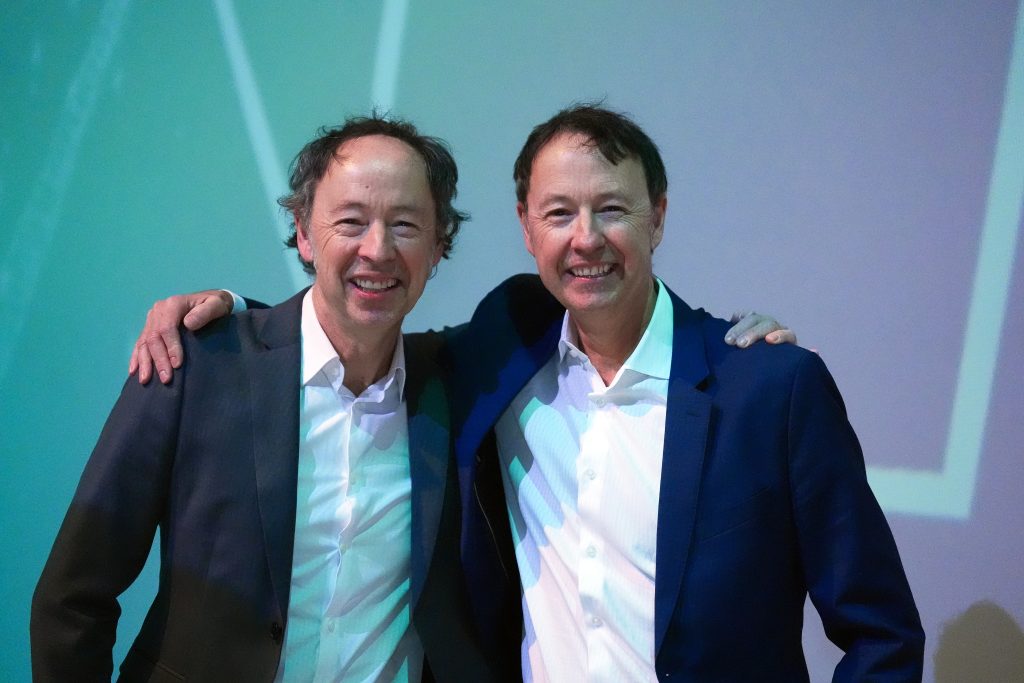 Zwei mittelalte Männer in Anzügen stehen nebeneinander, lächeln in die Kamera und haben die Arme umeinander gelegt.