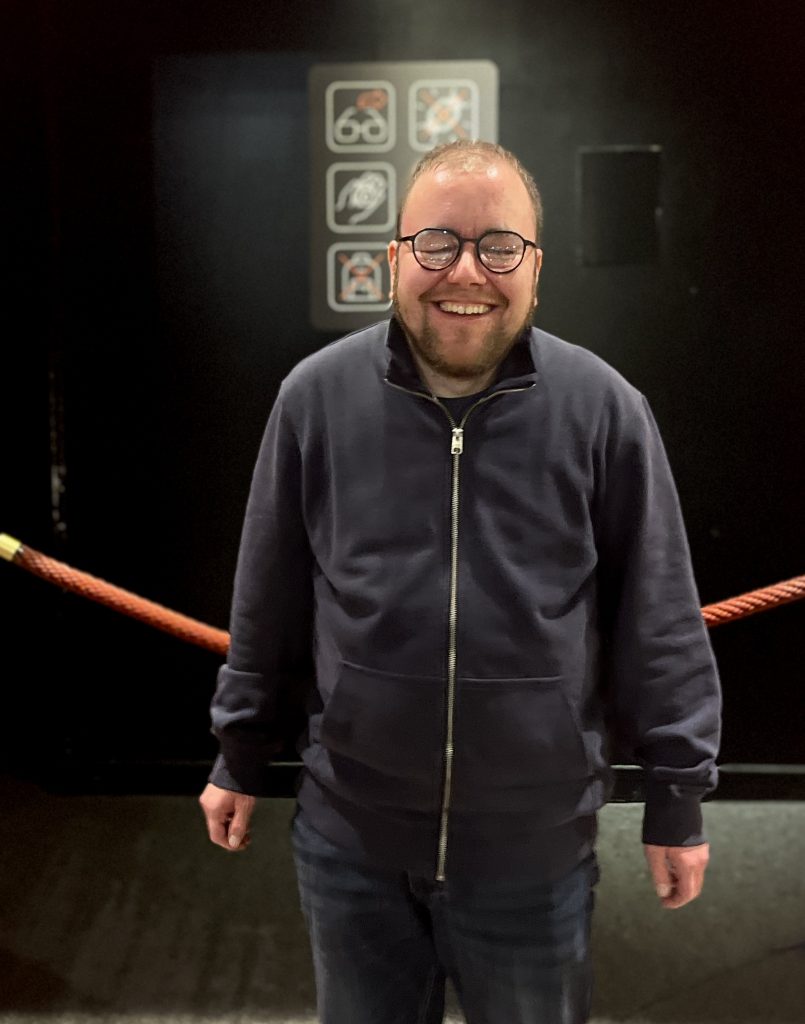 Ein Mann steht vor einem schwarzen Hintergrund, er lacht in die Kamera. Er trägt eine Brille, seine Augen sind geschlossen.