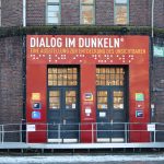 Wie fühlt es sich an, blind zu sein? Der Eingang des Dialoghauses mit dem Schriftzug Dialog im Dunkeln. Darunter nochmal in Blindenschrift. Darunter die Eingangstüren.