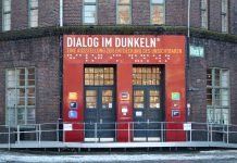 Wie fühlt es sich an, blind zu sein? Der Eingang des Dialoghauses mit dem Schriftzug Dialog im Dunkeln. Darunter nochmal in Blindenschrift. Darunter die Eingangstüren.