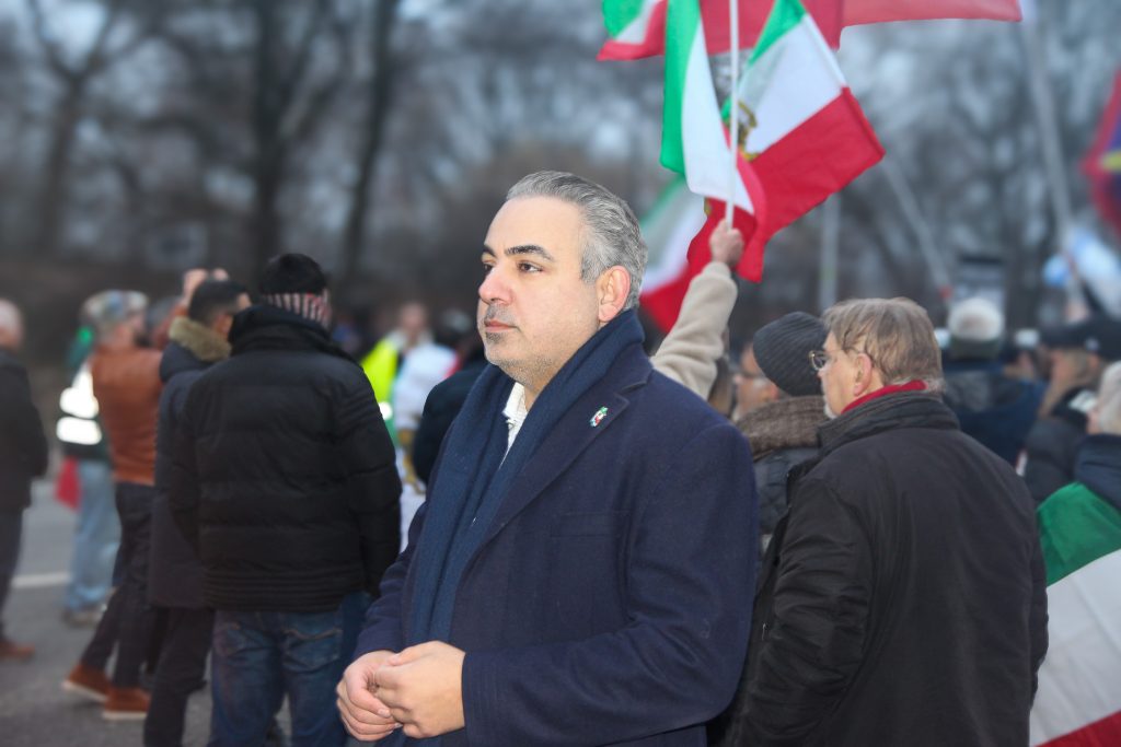 Ein Mann in einem blauen Mantel schaut ernst zur linken Seite. Dahinter sieht man Demonstrierende mit Iran-Flaggen.