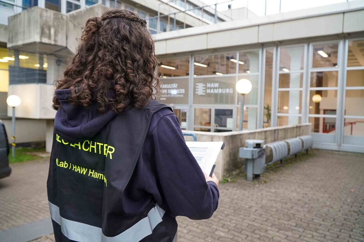Auf dem Foto sieht man eine junge Frau mit dunklen Haaren von hinten. Sie trägt eine schwarze Warnweste auf der Beobachter steht. Sie hält ein Klemmbrett mit Zetteln darauf und steht vor dem verglasten Eingang eines Gebäudes.