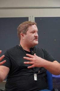 Auf dem Bild ist ein Mann mit braunen Haaren und einem Schnurrbart. Er scheint mitten im Gespräch und gestikuliert mit seinen Händen. An seinem T-Shirt Kragen ist ein kleines Mikrofon angesteckt.