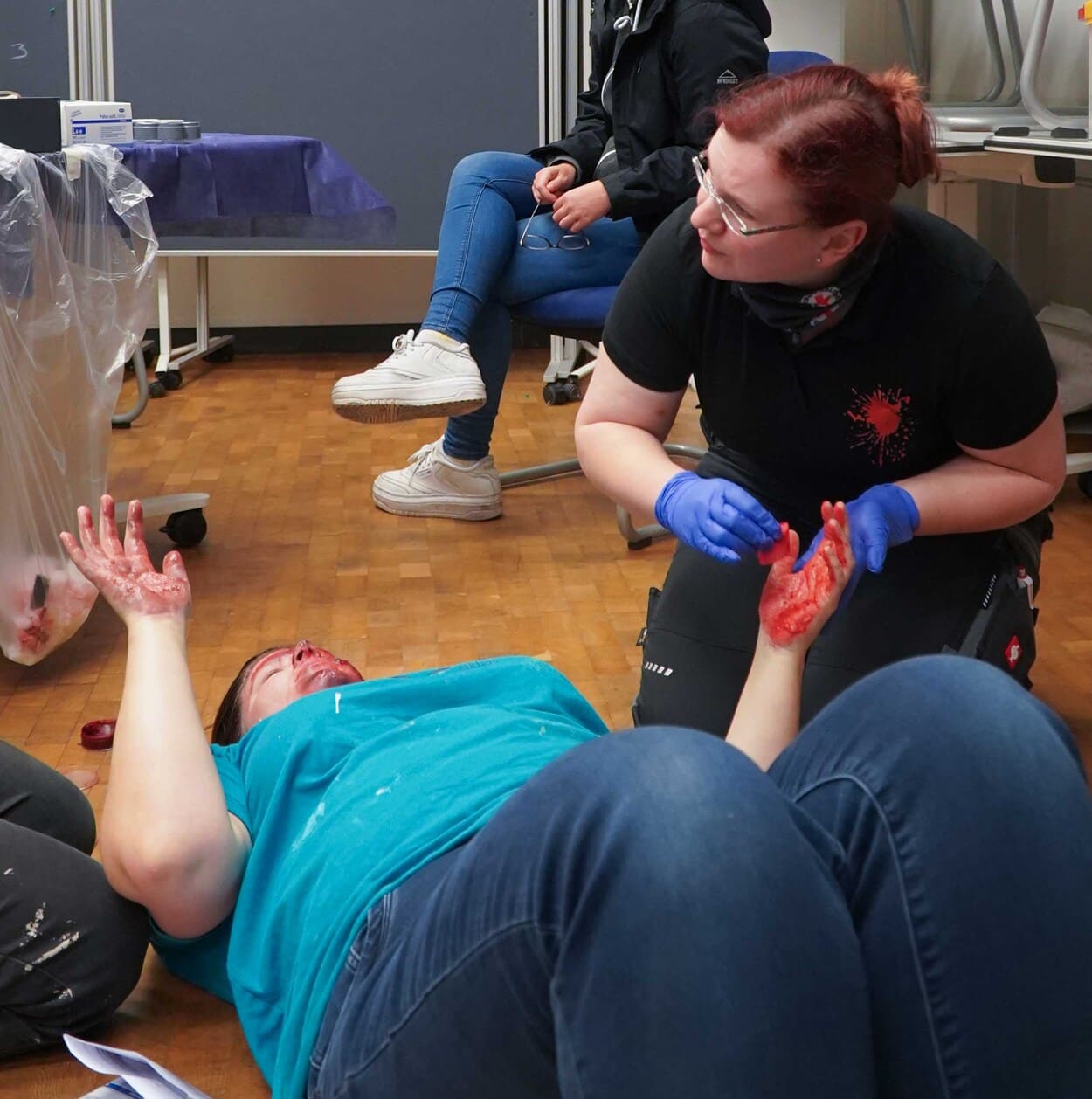 Eine Frau mit blauem T-Shirt liegt auf dem Boden. Eine zweite Frau mit blauen Einweghandschuhen pinselt ihr rotes Kunstblut auf die Hände.