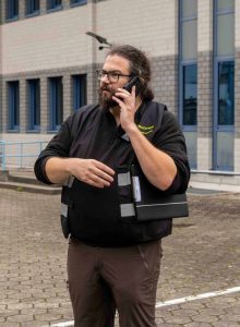 Auf dem Bild ist ein Mann mit Brille, dunkelbraunen Haaren und Bart zu sehen, der gerade telefoniert. Unter dem Arm hat er ein Klemmbrett. Im Hintergrund ist ein helles Gebäude mit blauen Fenstern zu sehen.