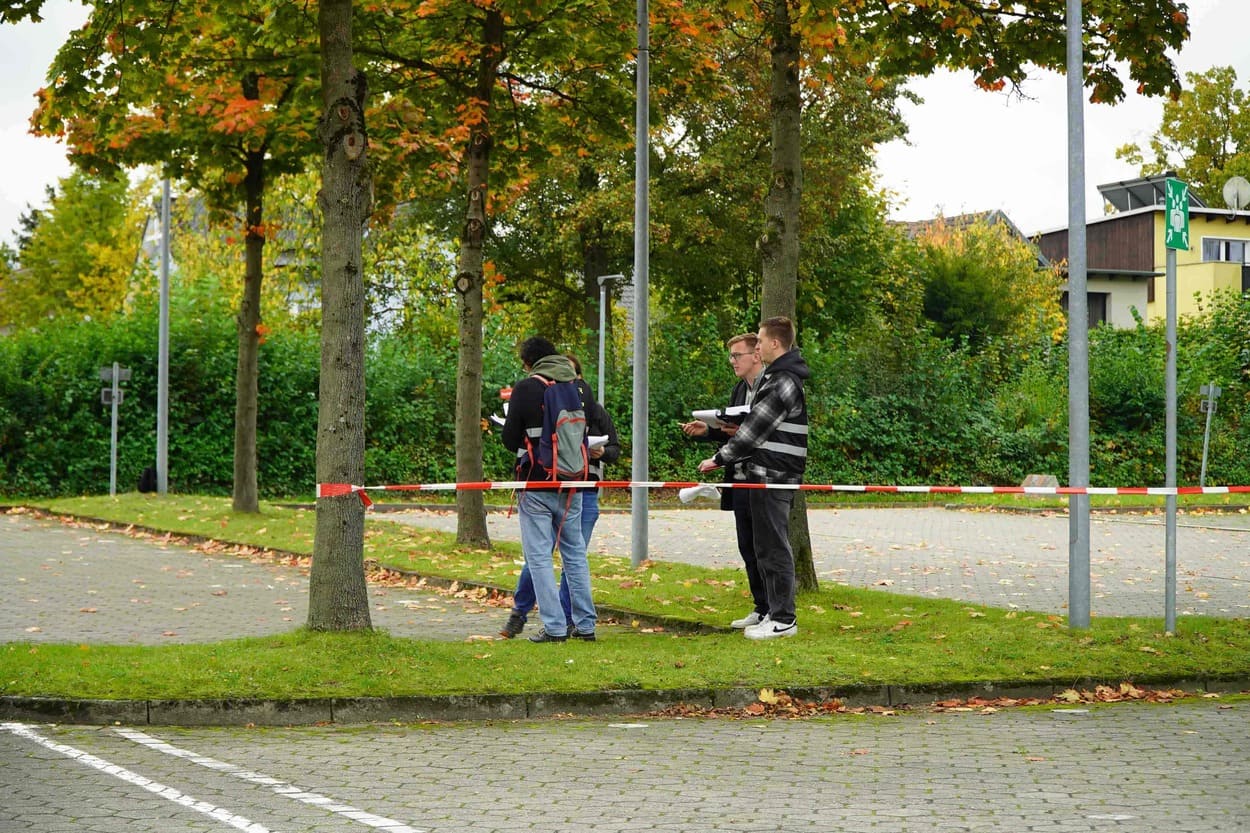 Auf dem Bild sieht man von ca. 20 Metern Entfernung vier junge Personen, die auf einer kleinen Grünfläche auf einem Parkplatz stehen. Neben ihnen steht ein Baum. Daran ist ein Absperrband befestigt.