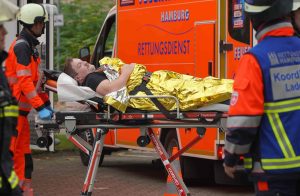 Ein Darsteller mit Schnurrbart liegt auf einer Trage und wird zu einem Rettungswagen gefahren. Er ist in eine goldene Wärmedecke gehüllt und an der Trage festgegurtet. Um ihn herum stehen Einsatzkräfte der Feuerwehr und des Rettungsdienstes.