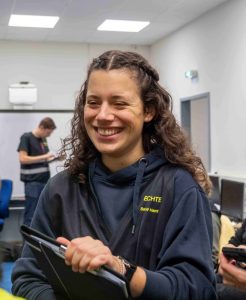 Auf dem Bild sieht man eine junge Frau mit dunklen, offenen Haaren. Sie hat ein Klemmbrett in der Hand und lacht in Richtung der Kamera.
