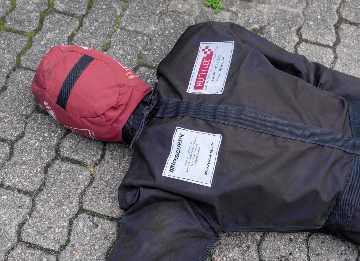 Man sieht den Oberkörper einer lebensgroßen Stoffpuppe, die auf dem asphaltierten Boden liegt. Der Kopf ist aus rotem Material. Die Puppe trägt ein braunes Oberteil mit zwei weißen Aufnähern.