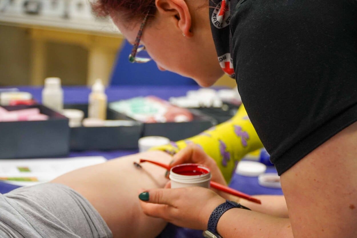 Eine Frau malt mit einem Pinsel und roter Farbe auf einem Bein. Sie schminkt darauf eine Verletzung.