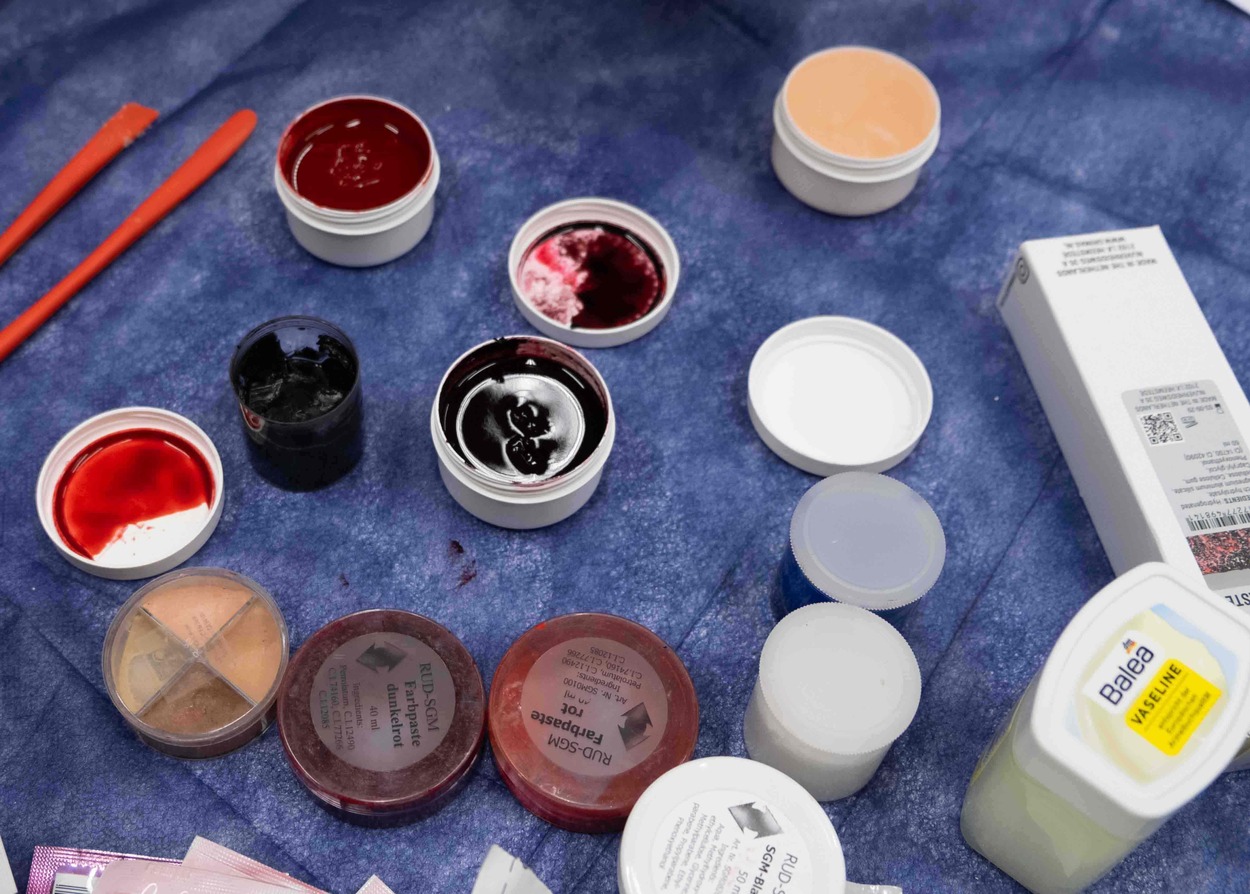 Auf einem Tisch mit einem blauen Vliestuch stehen etwa ein Dutzend kleine Tiegel mit verschiedenen Farbpasten. Auch ein Tiegel mit Vaseline steht auf dem Tisch.