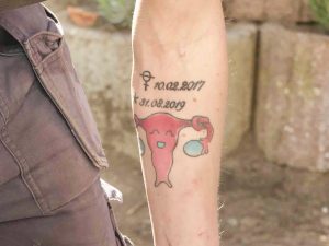 Ein buntes Tattoo auf dem Unterarm einer Frau zeigen zwei Daten, den 10.02.2017 und den 31.08.2019. Darunter eine Gebärmutter mit lachendem Gesicht.