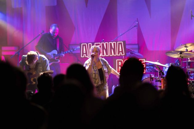 Eine Konzertb&uuml;hne mit lila Scheinwerferlicht. Durch die K&ouml;pfe zweier Personen aus dem Publikum ist eine S&auml;ngerin zu sehen.